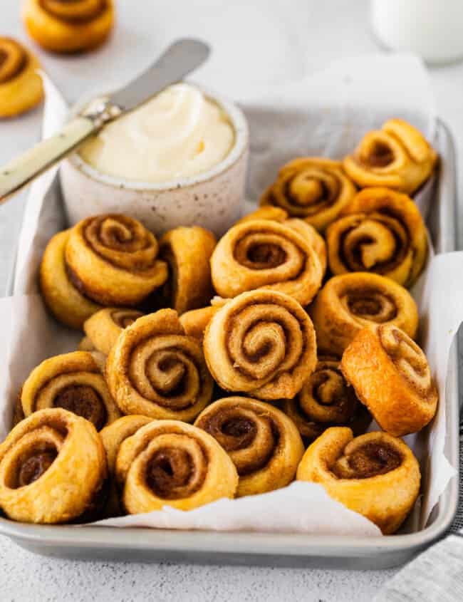 three-quarters view of mini cinnamon rolls in a serving tray with a small pot of frosting and a knife.