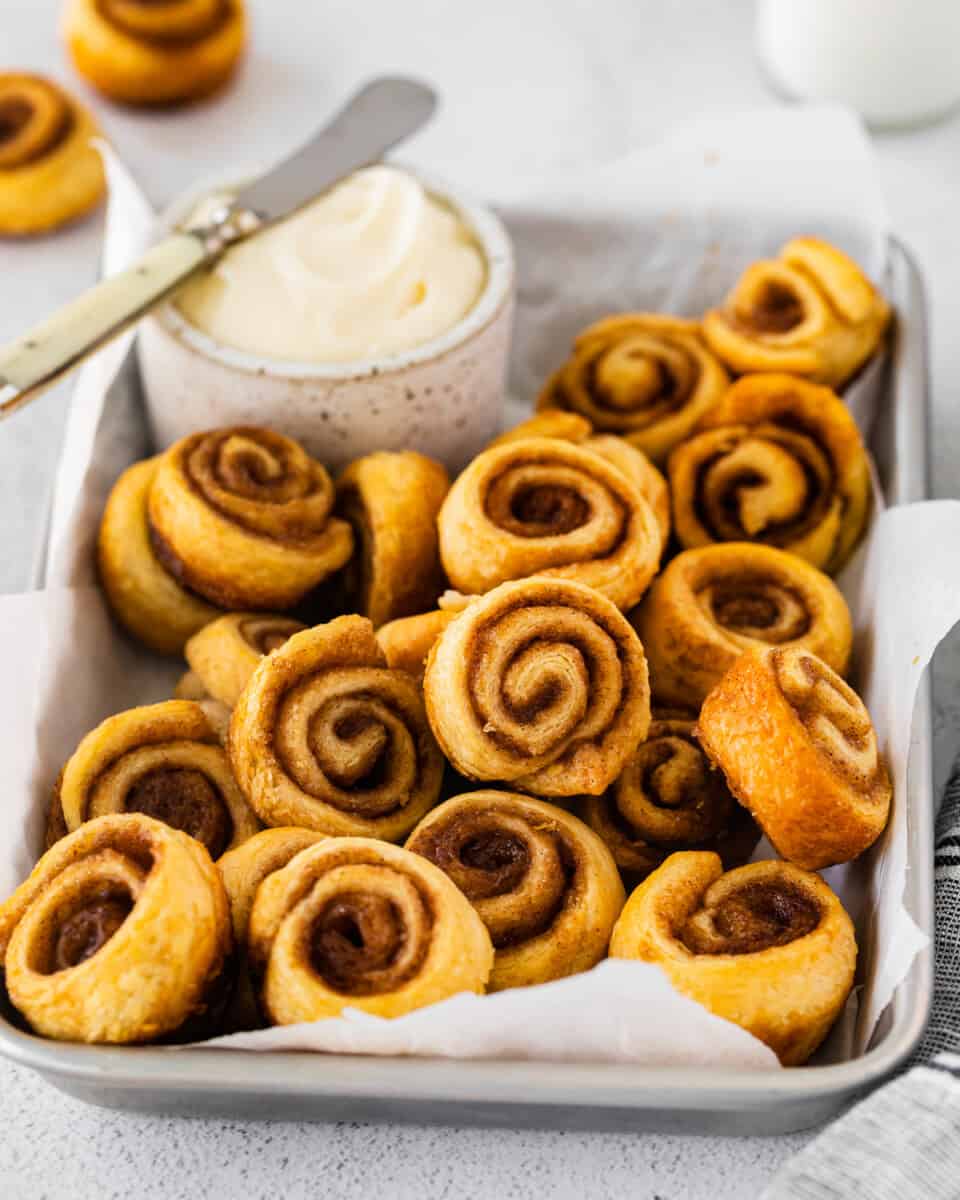 three-quarters view of mini cinnamon rolls in a serving tray with a small pot of frosting and a knife.