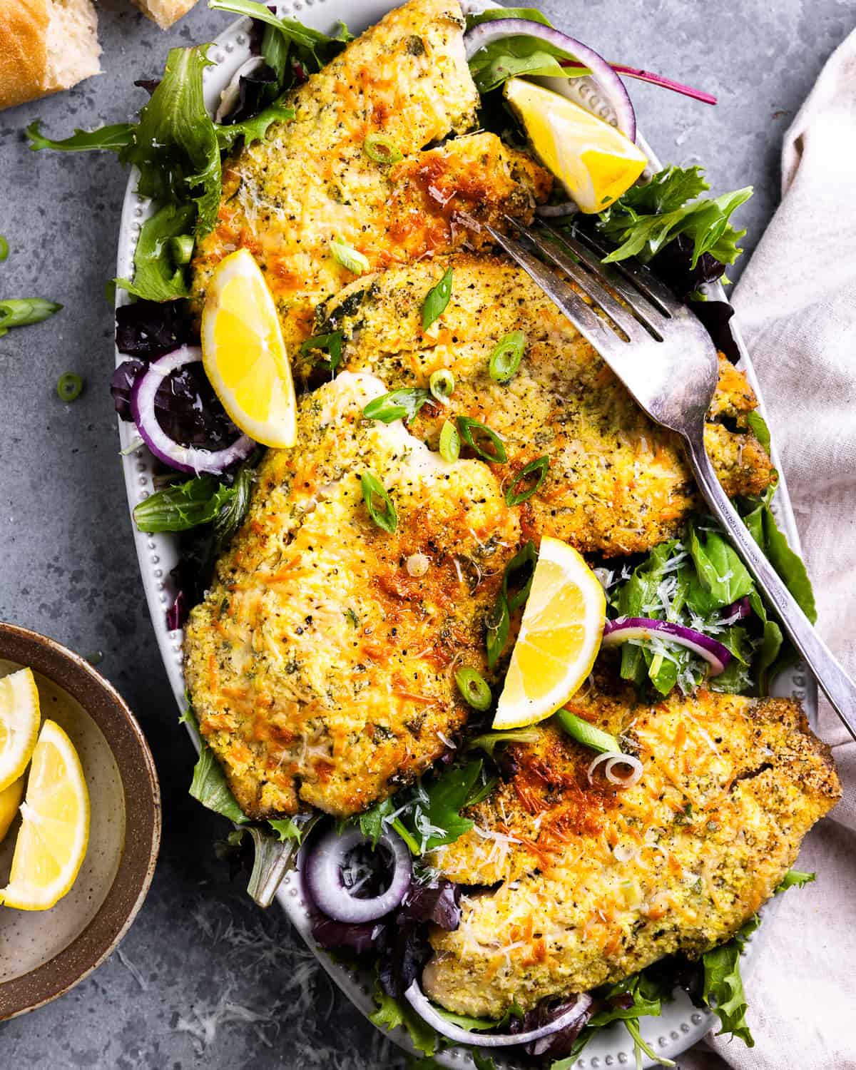 overhead view of parmesan crusted tilapia filets on an oval platter with salad greens, lemon wedges, and a fork.