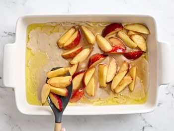 spooning sliced peaches over batter in a baking dish.