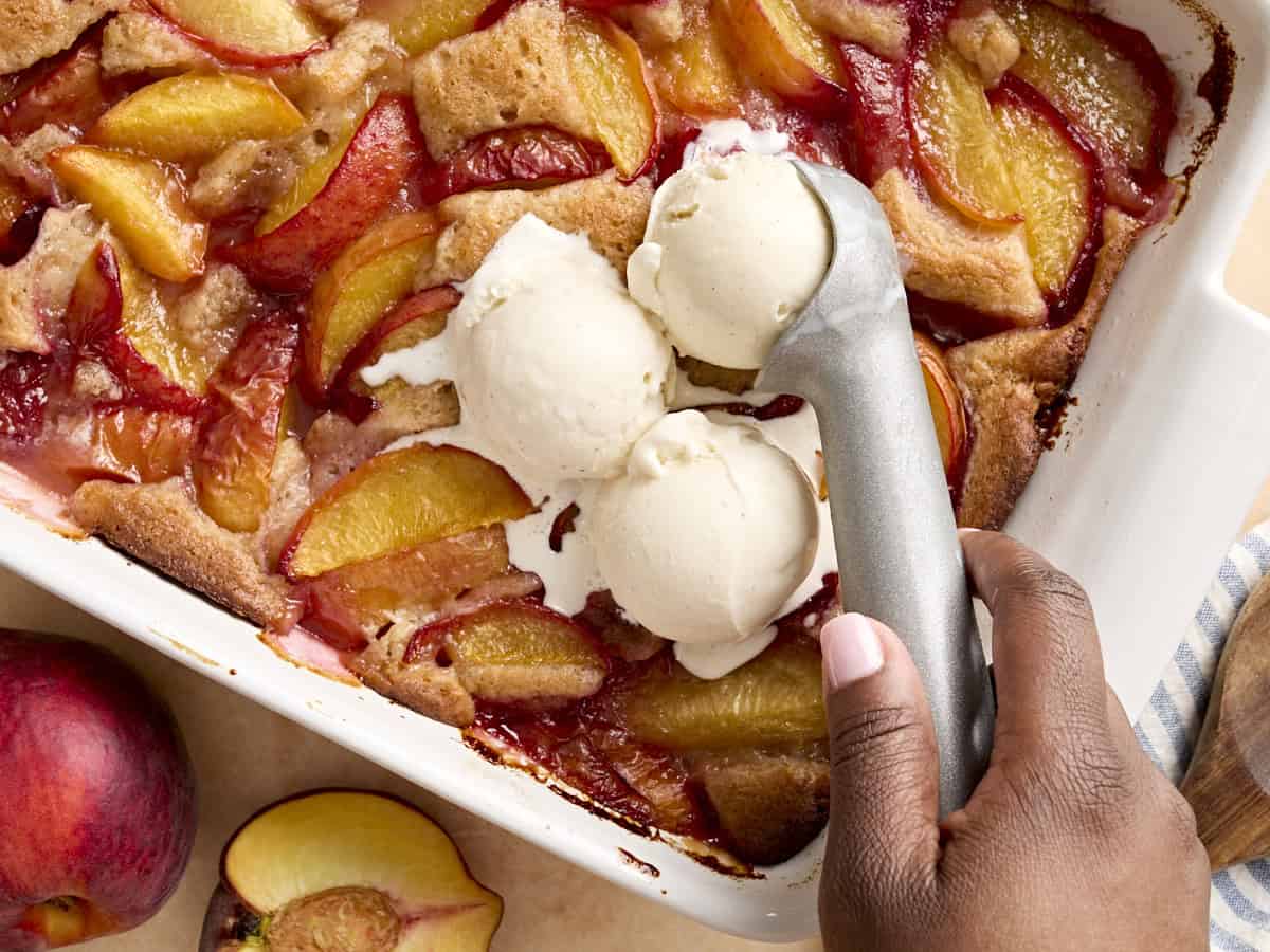 scooping ice cream over peach cobbler in a baking dish.