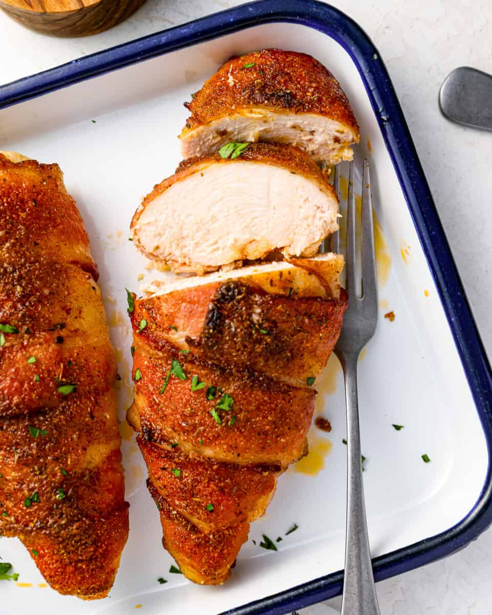 a sliced bacon wrapped chicken breast on a baking sheet with a fork.