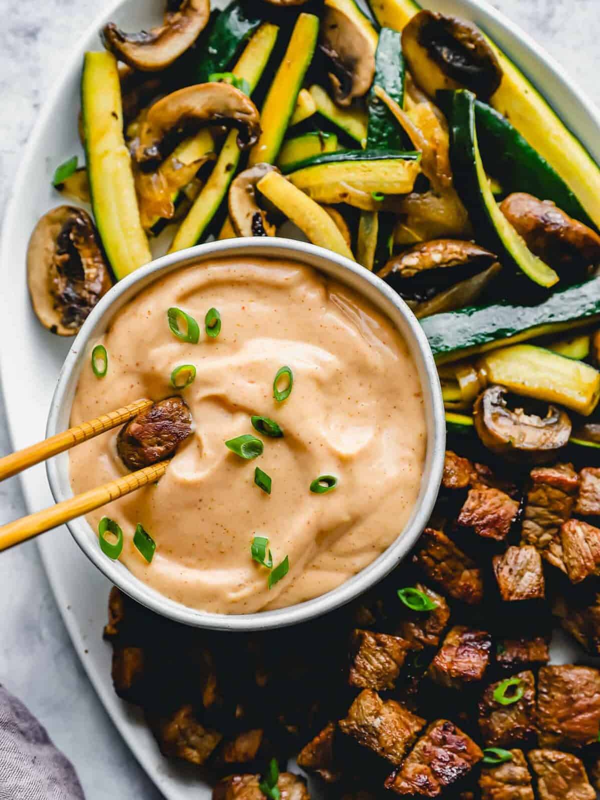 overhead view of a pair of chopsticks dipping a cube of steak into yum yum sauce in a white bowl.