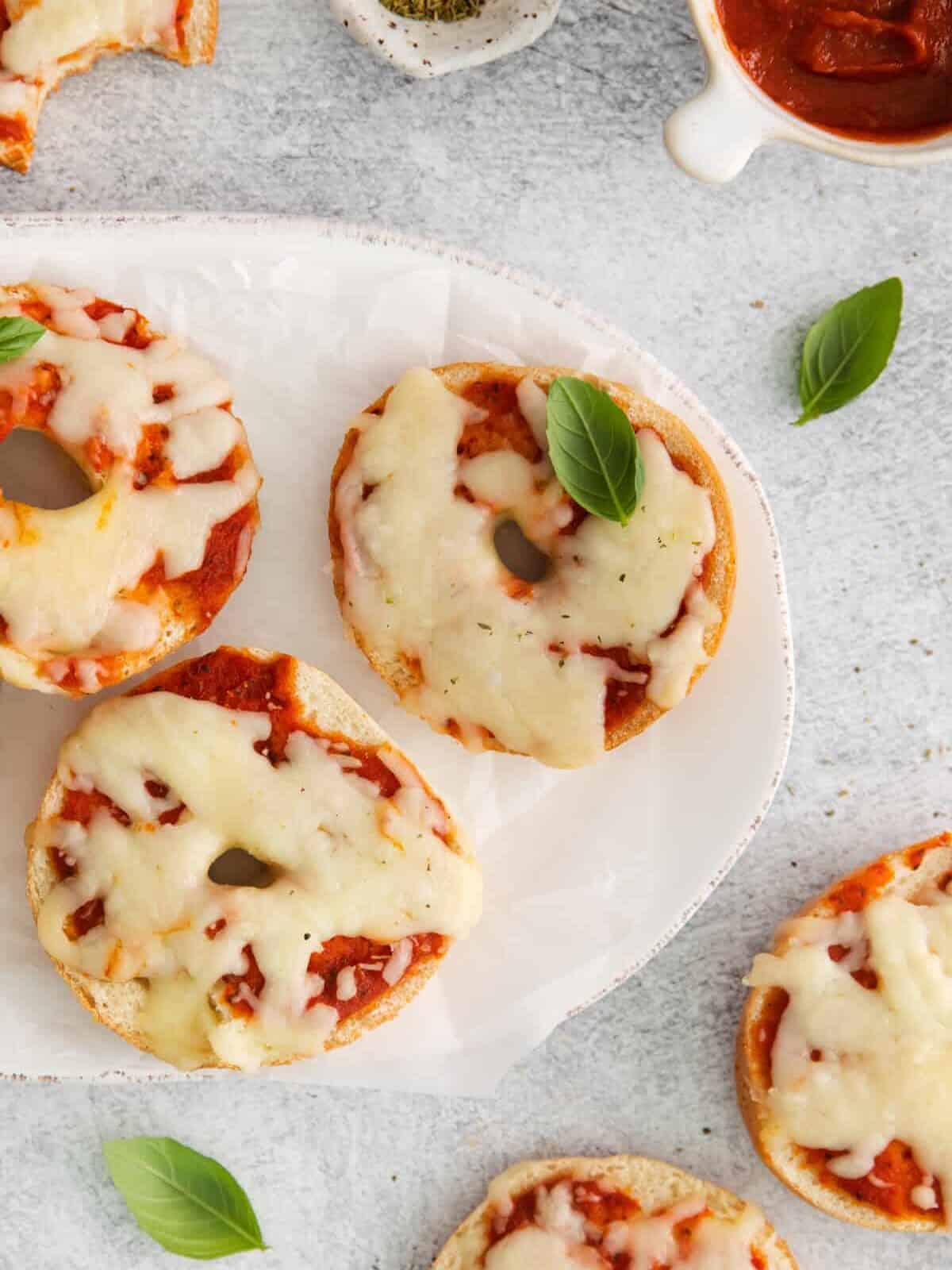 partial overhead view of 3 pizza bagels on an oval serving dish with more pizza bagels scattered around it.