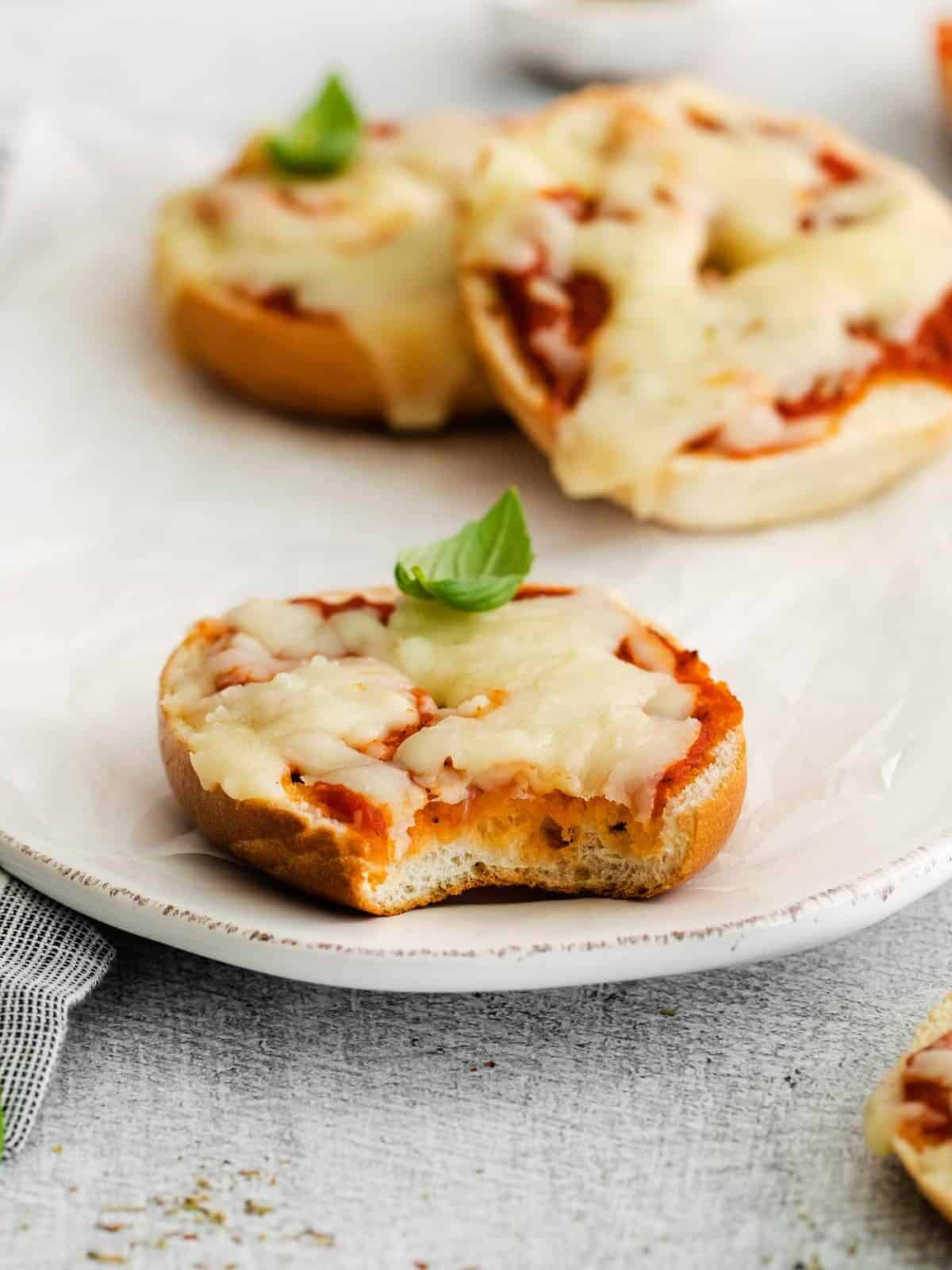 three-quarters view of a bitten homemade pizza bagel on a white oval serving dish topped with a basil leaf.