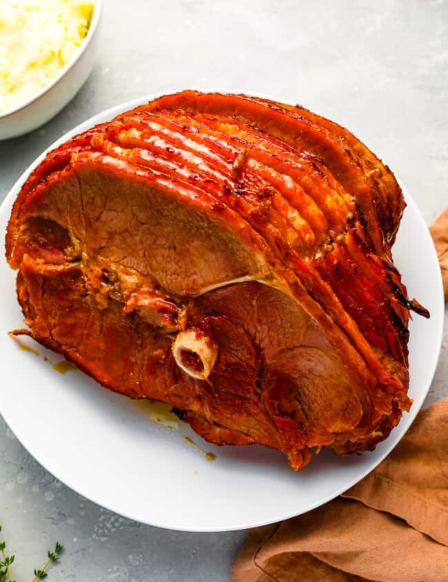 A roasted ham on a plate next to a bowl of mashed potatoes.