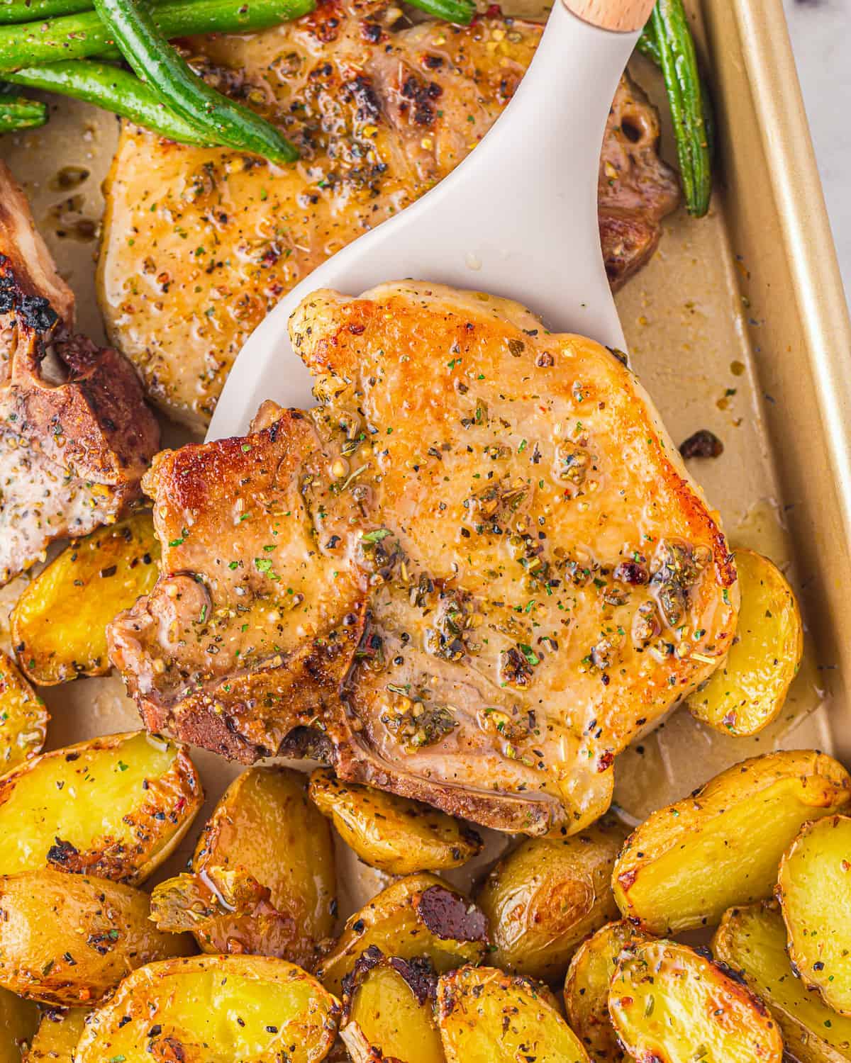 Spatula lifting up a pork chops from a pan of potatoes.