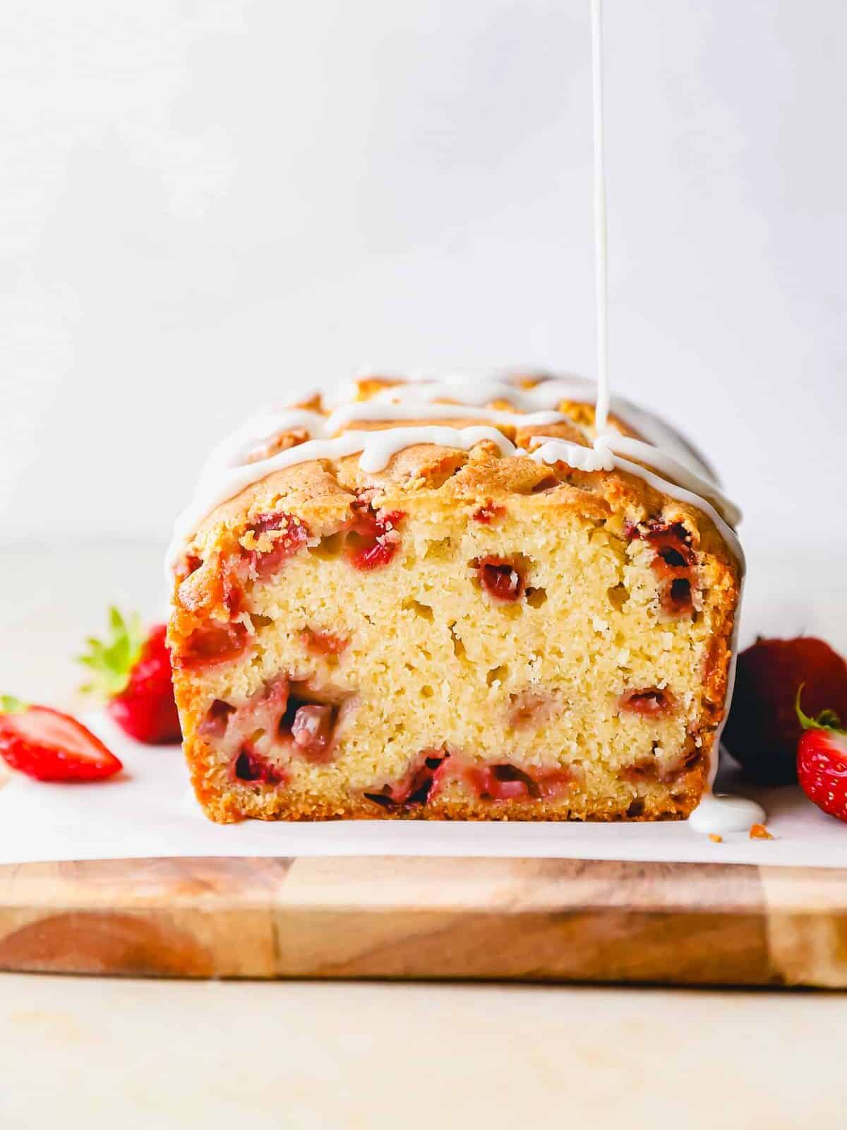 side view of glaze being drizzled over strawberry bread on a wooden cutting board that has been sliced open to show the interior.