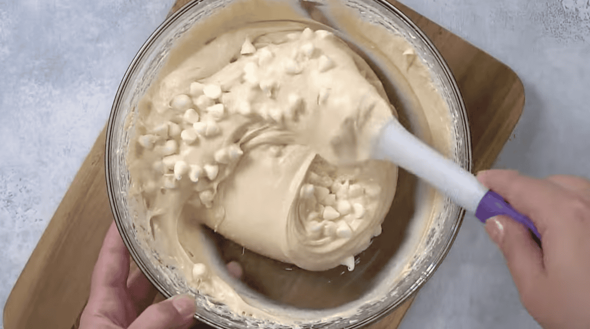 folding white chocolate chips into caramel apple dip in a glass bowl with a rubber spatula.