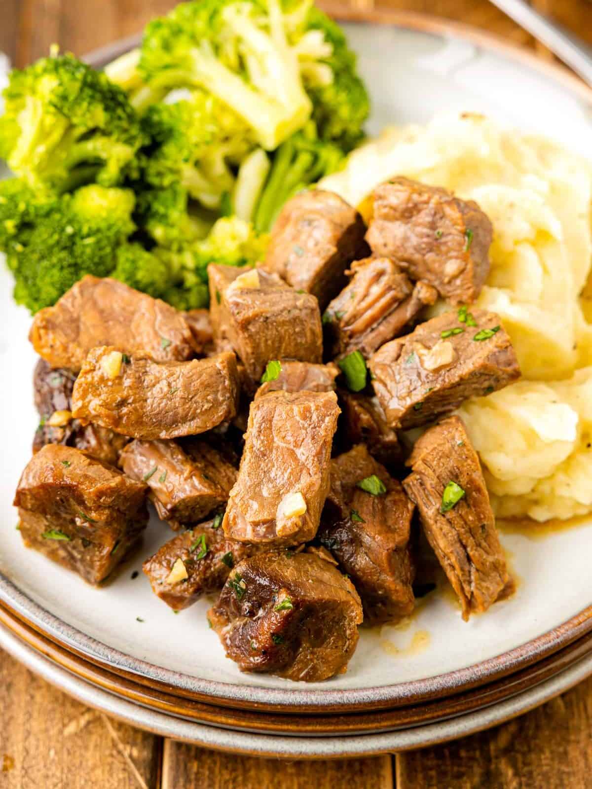 crockpot steak bites on a white plate with mashed potatoes and broccoli.