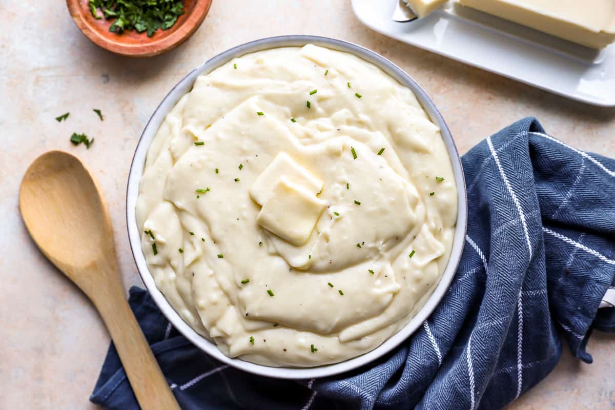 A bowl of cheesy pommes aligot fondue mashed potatoes on a table with a dish towel, wooden spoon, and tray of butter.