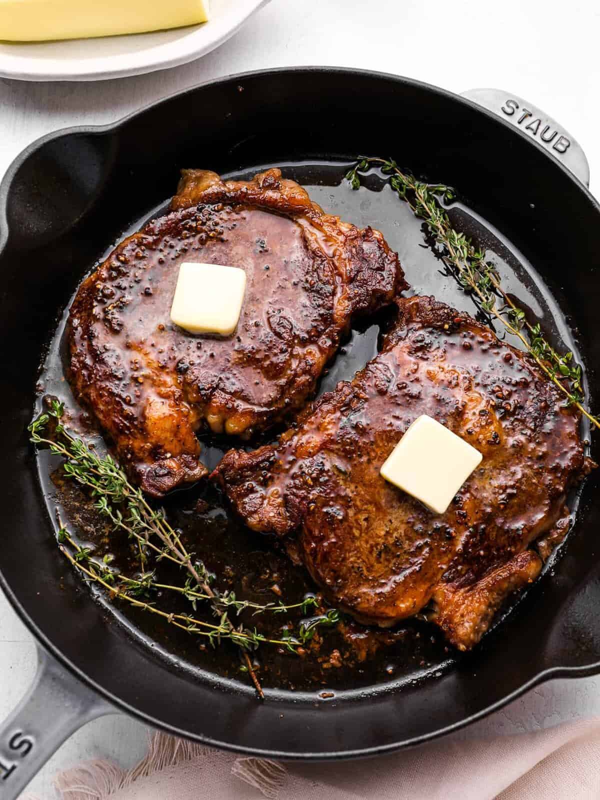 overhead view of oven baked steaks in a cast iron pan, topped with a pat of butter and thyme.