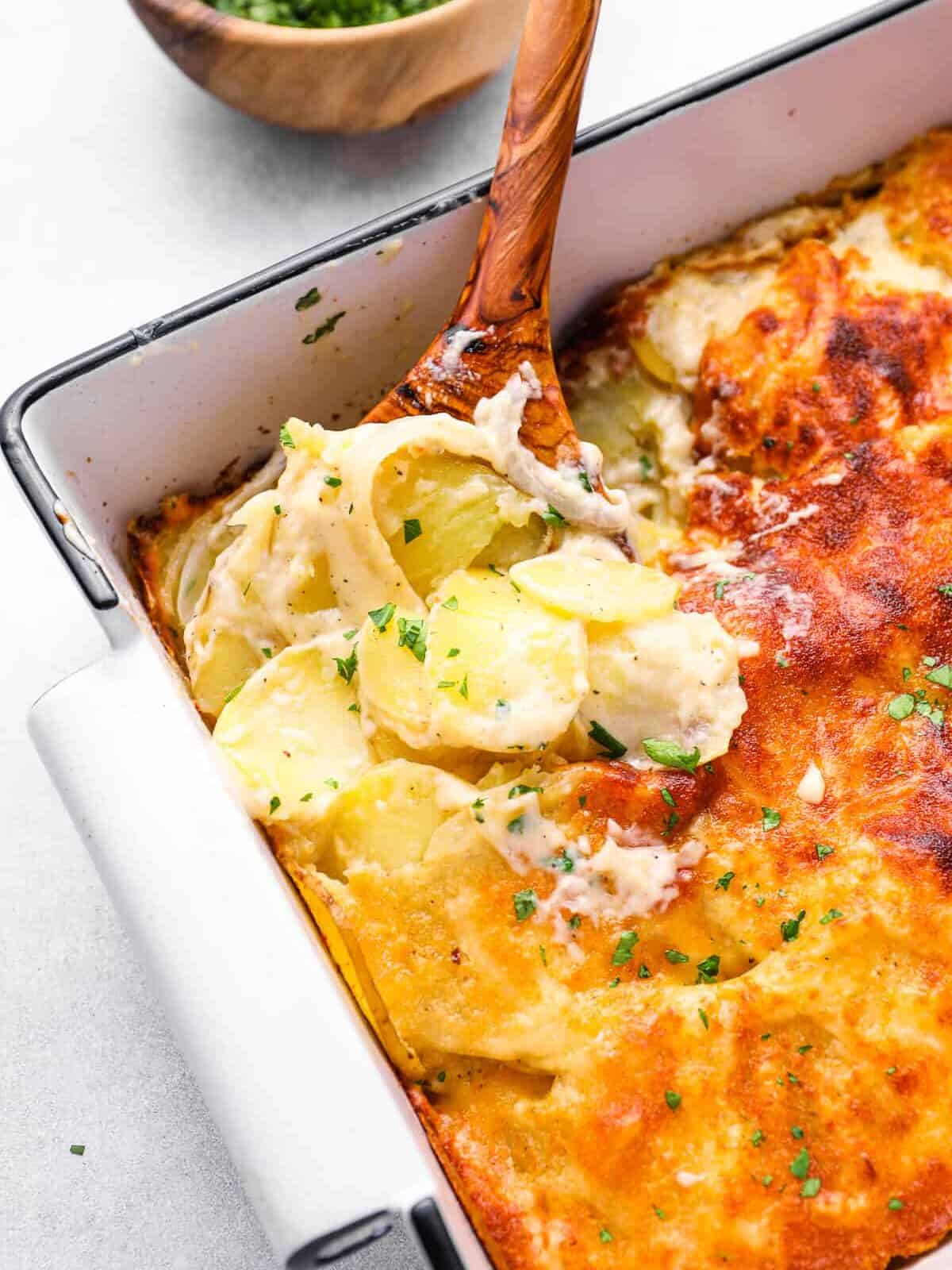 a wooden spoon scooping au gratin potatoes out of a 9x13 baking pan.