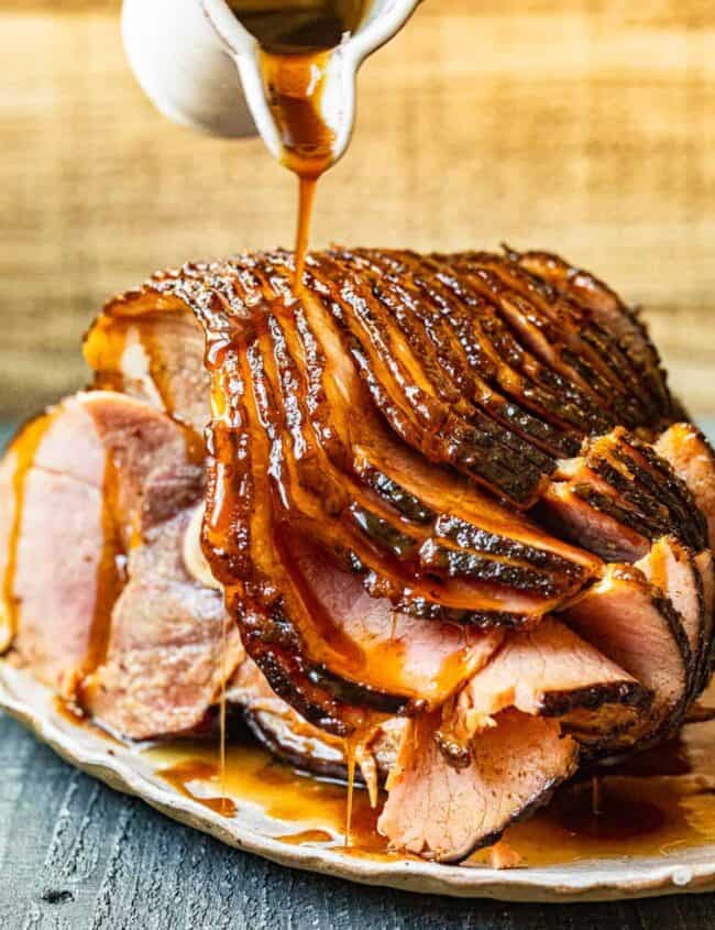 hand pouring glaze onto honey baked ham on a white serving platter