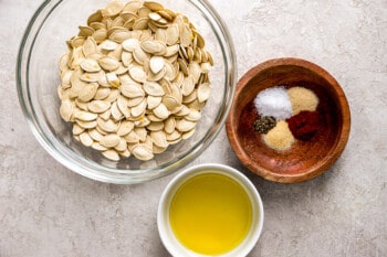 Pumpkin seeds in a bowl next to a bowl of oil and salt.