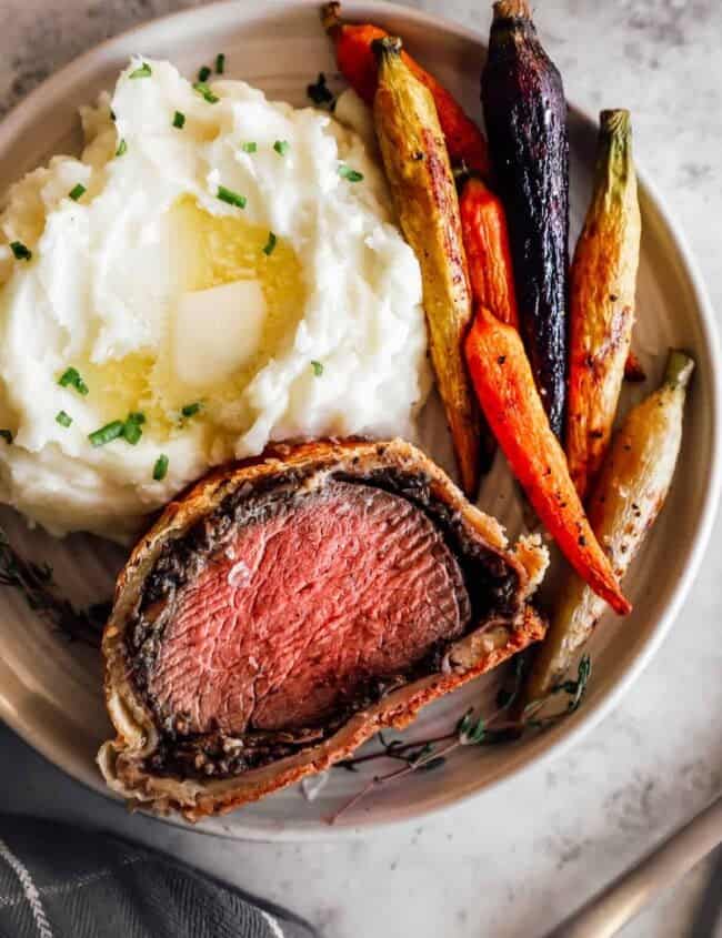 a slice of beef wellington on a white plate with carrots and mashed potatoes.