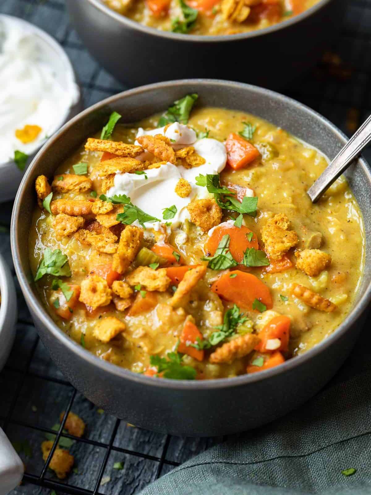 three-quarters view of a serving of mulligatawny soup in a gray bowl with a spoon, topped with crumbled crackers and a dollop of yogurt.