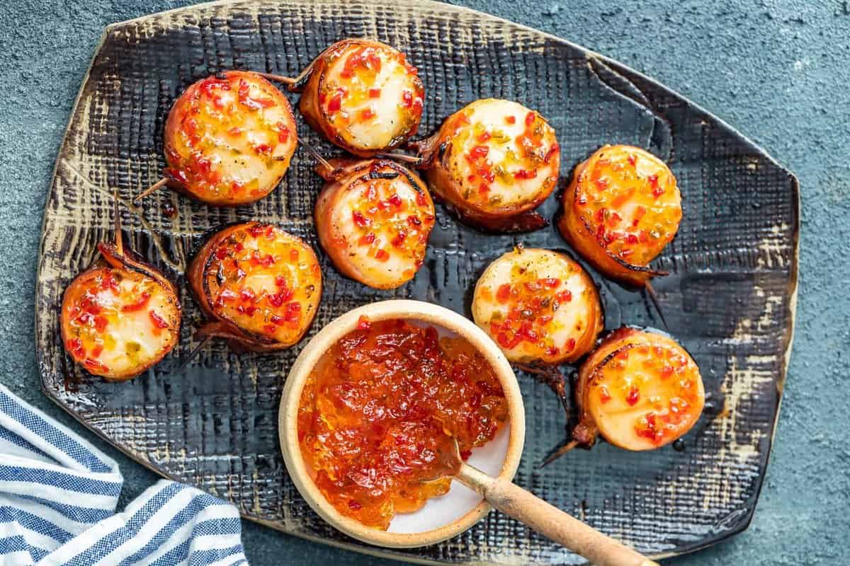 a serving platter with bacon wrapped scallops and a bowl of hot pepper jelly