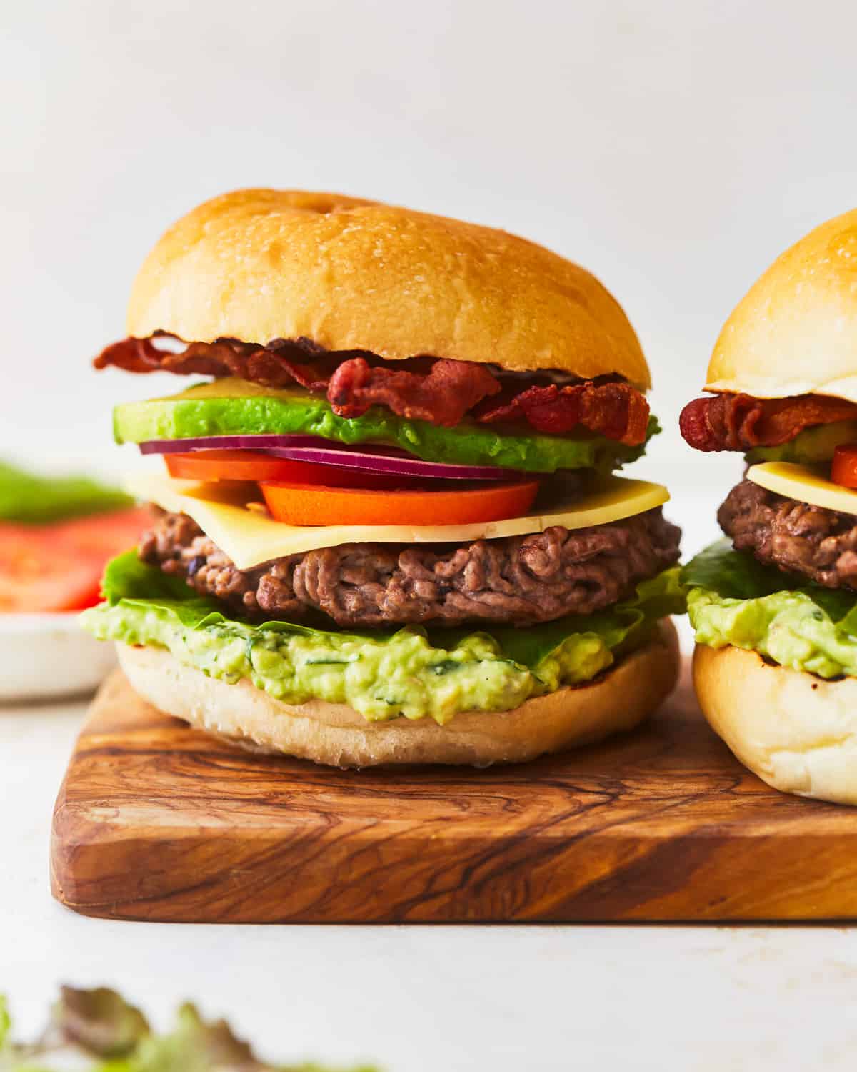 Two burgers with avocado and bacon on a wooden cutting board.