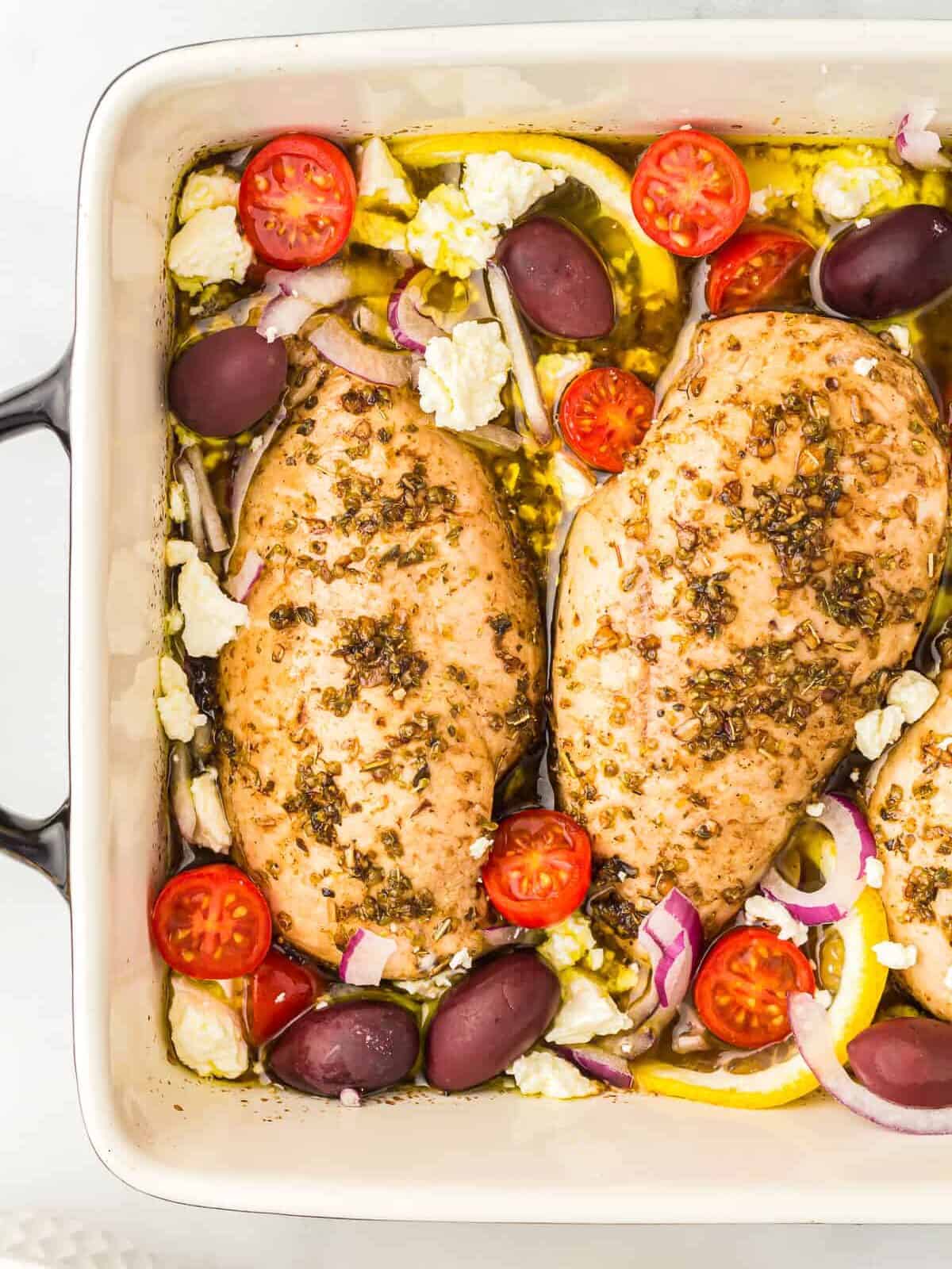 partial overhead view of baked greek chicken with tomatoes, red onions, feta, and olives in a rectangular baking pan.