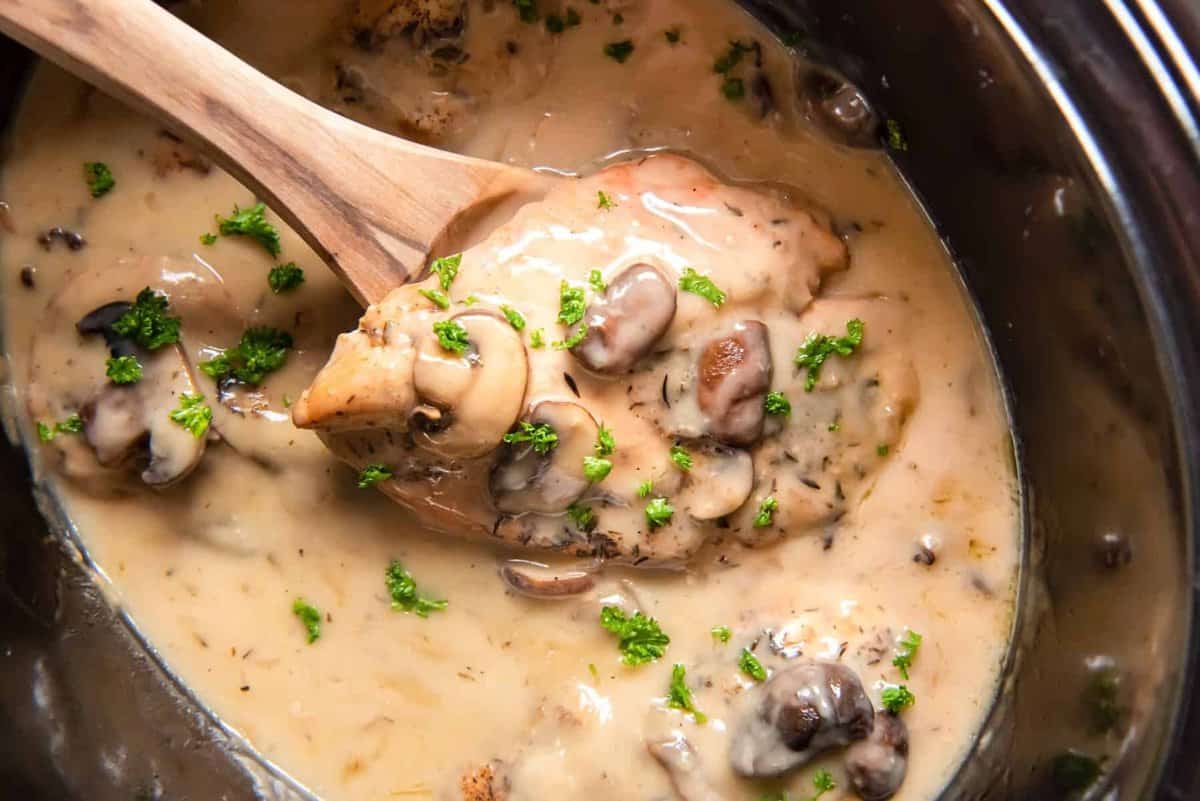 a wooden spoon lifting a chicken breast out of crockpot chicken marsala.