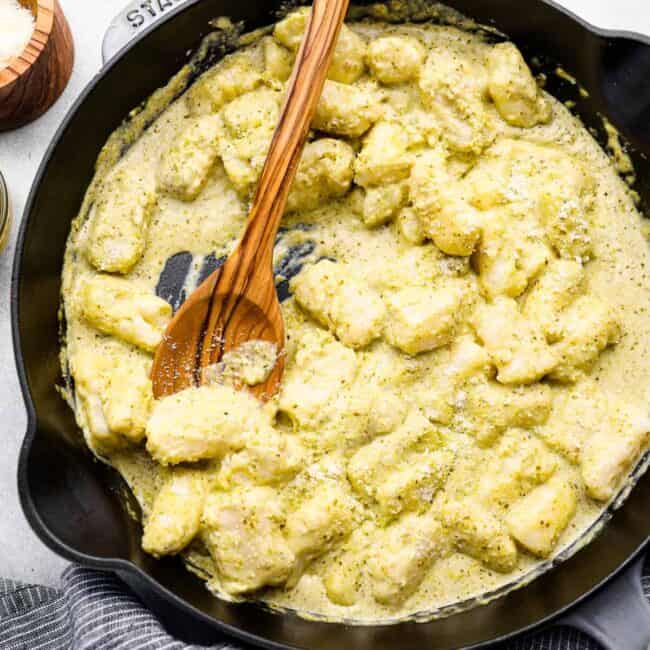 overhead view of gnocchi in pesto cream sauce in a cast iron skillet with a wooden spoon.