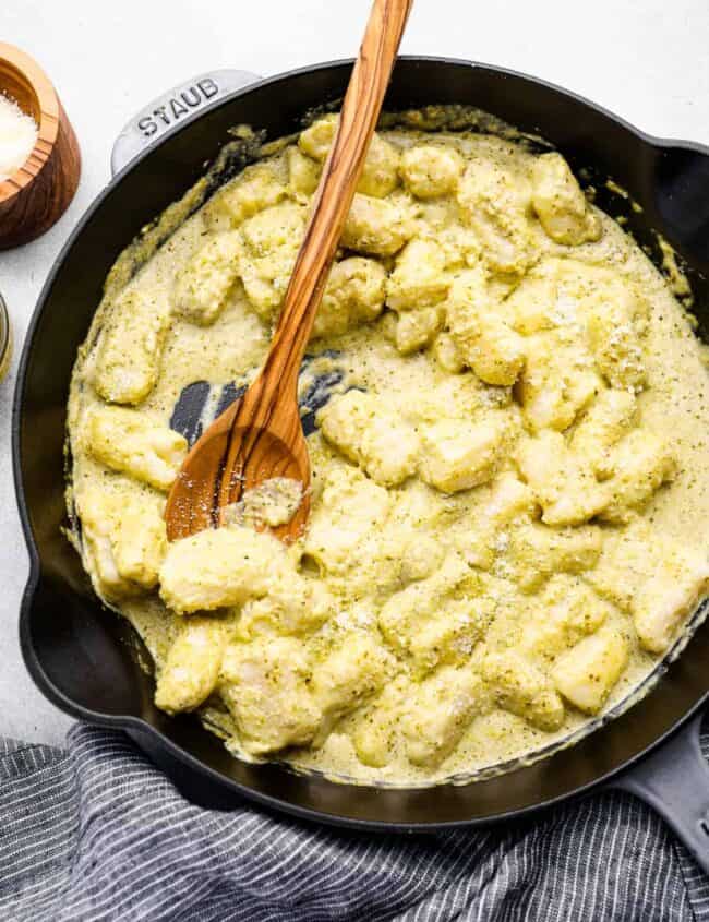 overhead view of gnocchi in pesto cream sauce in a cast iron skillet with a wooden spoon.