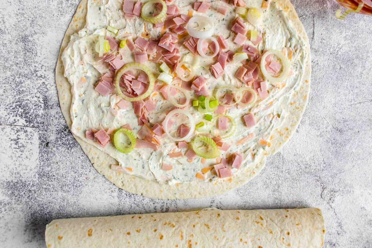 tortilla laid flat with cream cheese, ham, cheese, ranch, and green onions spread out on top.