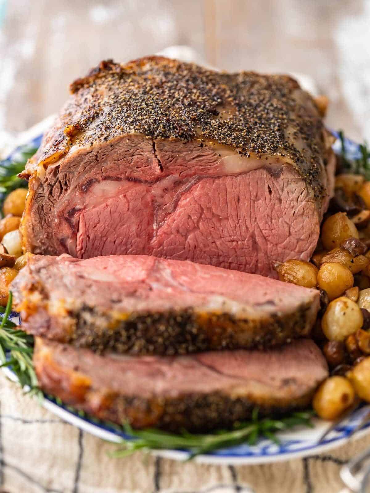 prime rib roast on a serving platter with potatoes, with a slice cut away. viewed from the front to reveal a red-pink medium-rare interior.