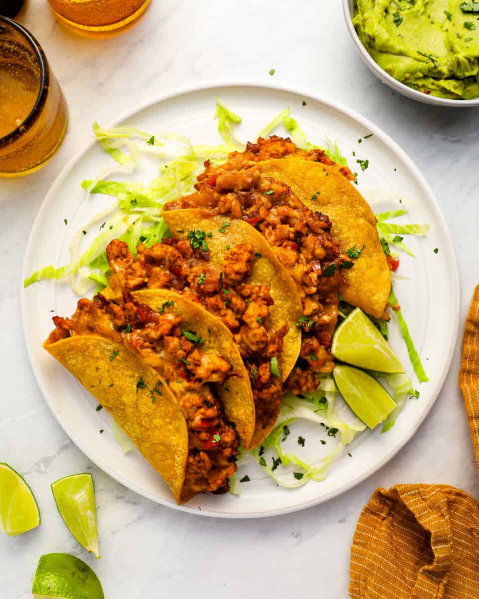 overhead view of 4 crispy chicken tacos on a white plate with lime wedges.