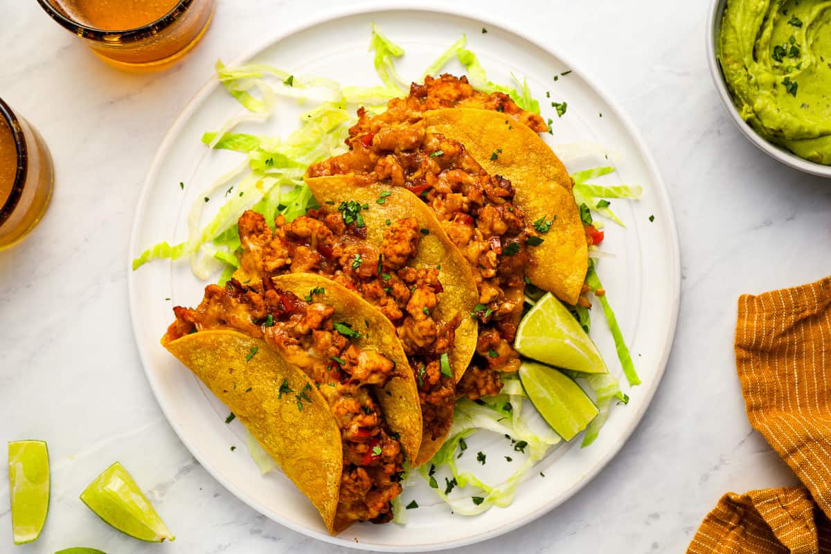 overhead view of 4 crispy chicken tacos on a white plate with lime wedges.