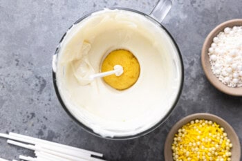 a lemon cake pop on a stick, dipped into a bowl of melted white chocolate.