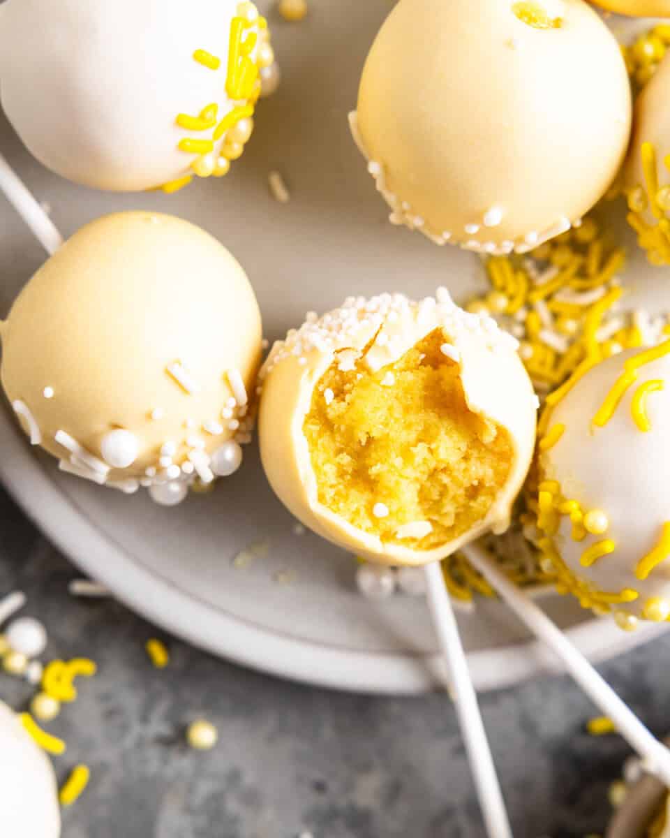 Close up on cake pops sitting on a plate; one has a bite taken out of it to reveal the lemon cake beneath the melted chocolate coating.