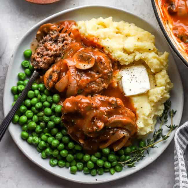 2 hamburger steaks on a white plate with peas, mashed potatoes, and a fork.