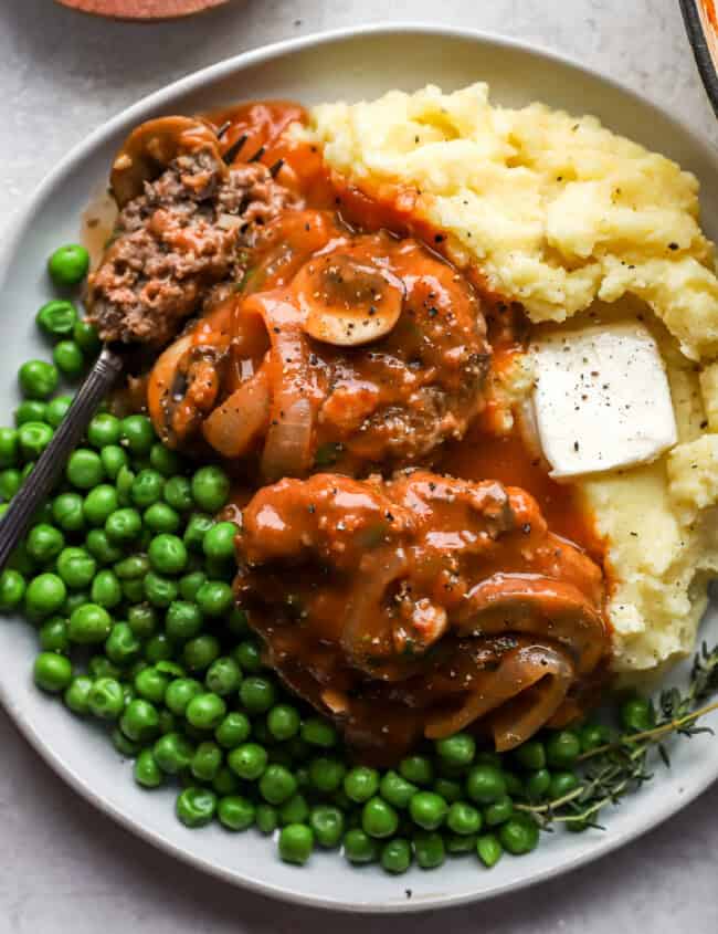 2 hamburger steaks on a white plate with peas, mashed potatoes, and a fork.