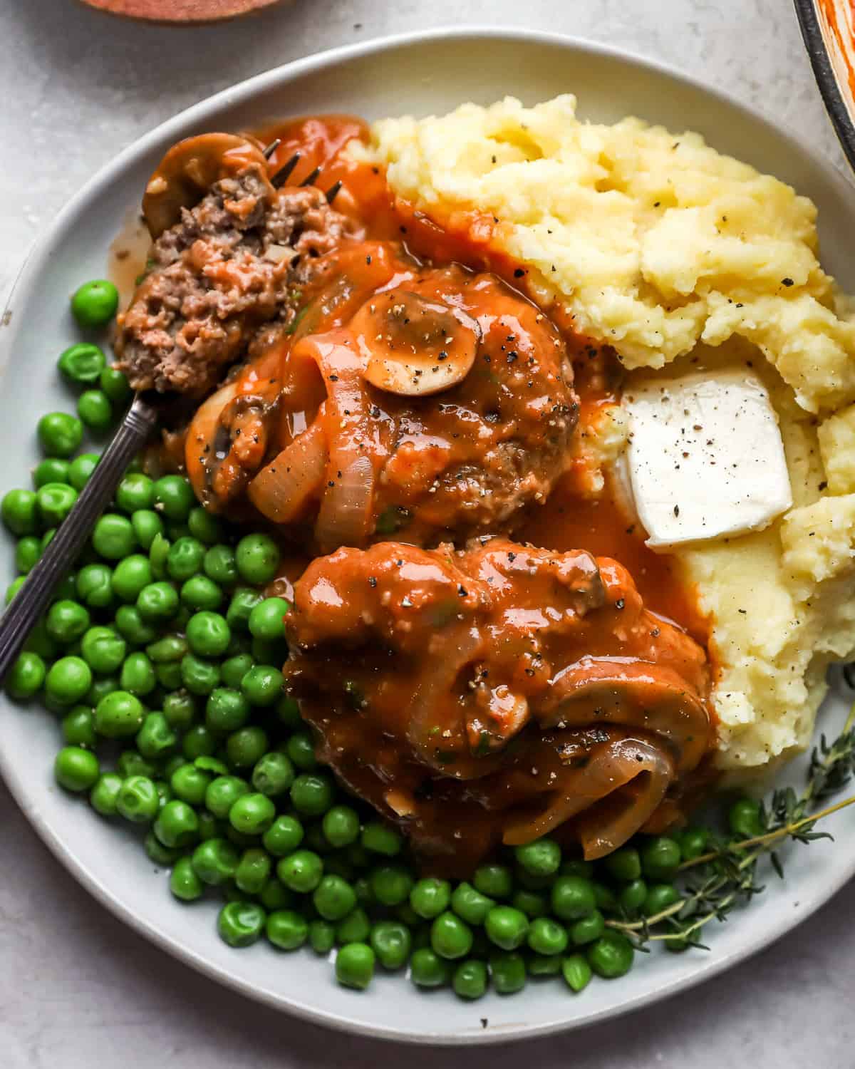 2 hamburger steaks on a white plate with peas, mashed potatoes, and a fork.