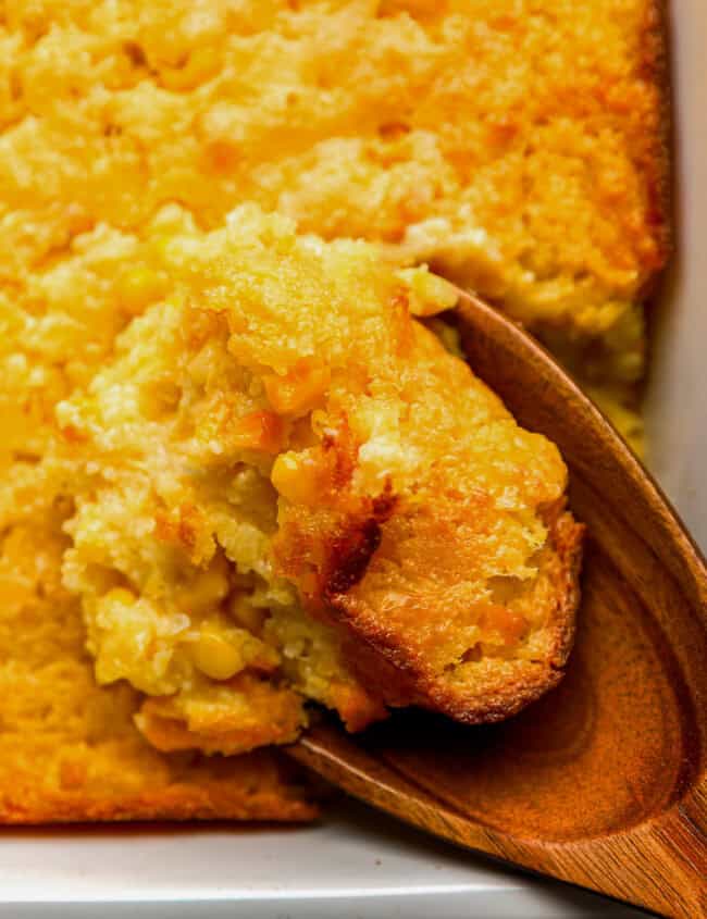 close up of a wooden spoon lifting a scoop of Jiffy corn casserole from a baking dish.