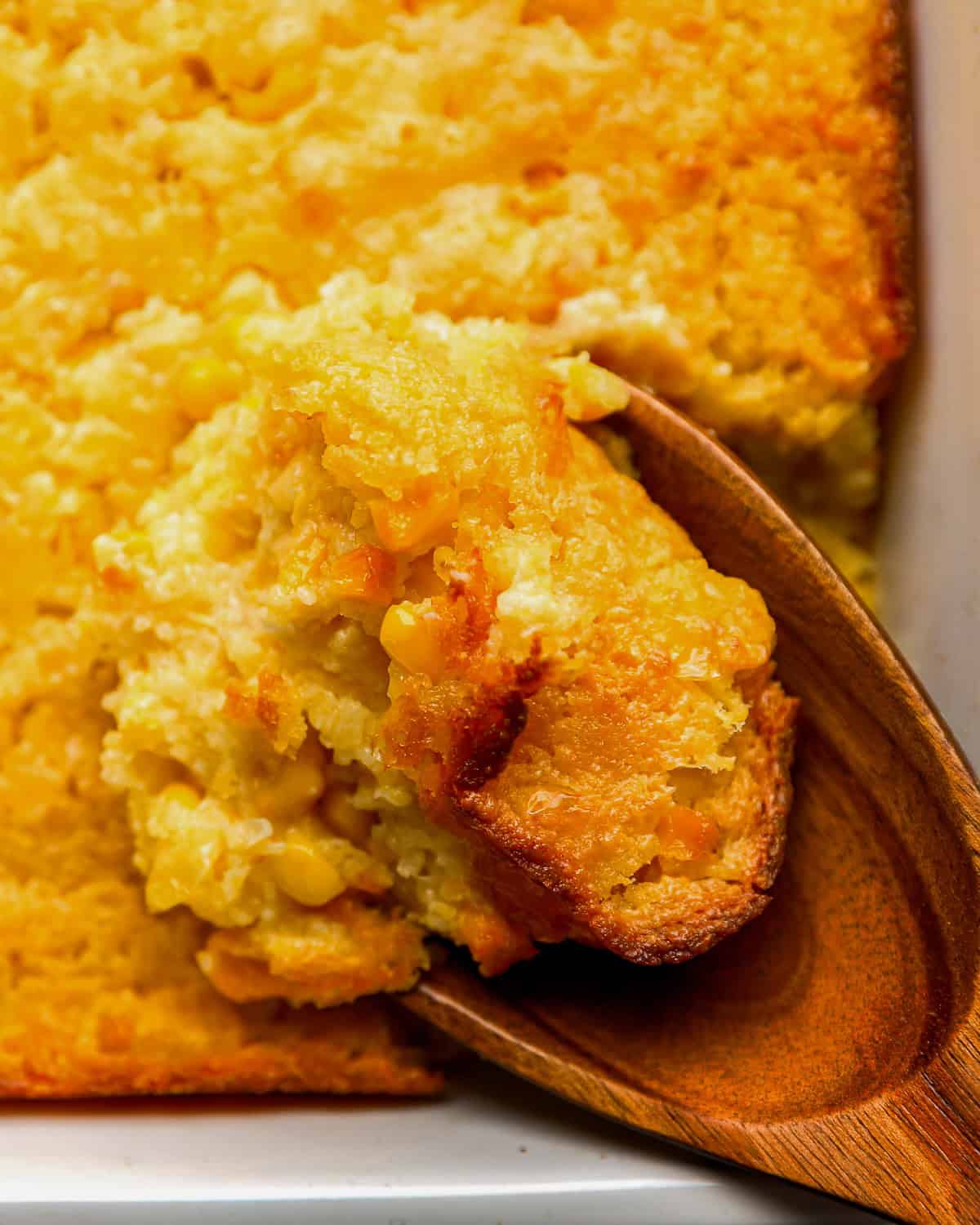 close up of a wooden spoon lifting a scoop of Jiffy corn casserole from a baking dish.