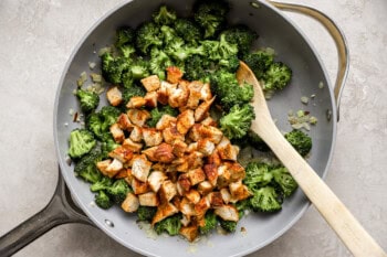 Broccoli florets and cubes of chicken in a skillet with a wooden spoon.