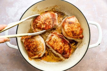 pulling a seared pork chop from a dutch oven with tongs.