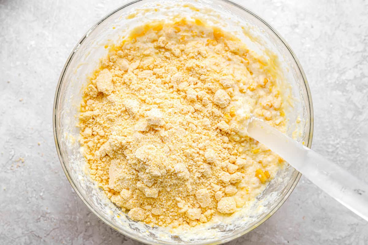 Jiffy corn muffin mix poured over corn casserole ingredients in a glass bowl with a spoon.