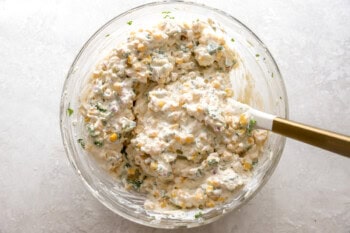 folding mexican street corn dip in a glass bowl with a rubber spatula.