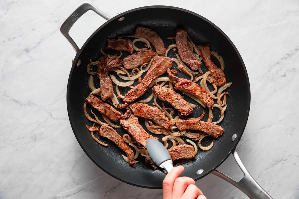 Hand using a tong to flip over a piece of steak in a skillet full of steak and onions.