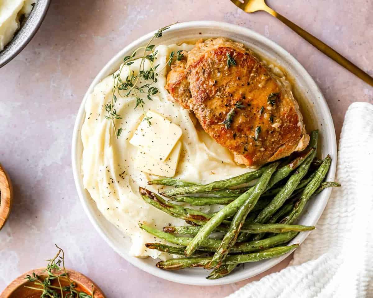 overhead view of a crockpot pork chop on a white plate with green beans and mashed potatoes.