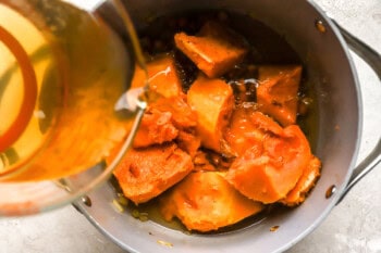 pouring chicken broth over butternut squash chunks and sauteed vegetables in a large pot.