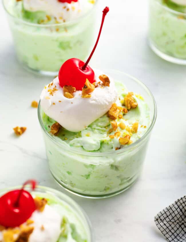 a serving of watergate salad in a glass dish with whipped cream, nuts, and a cherry on top.